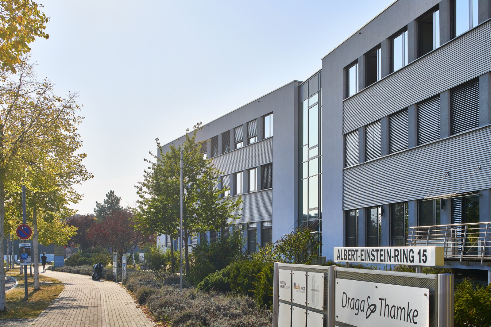 Bürogebäude neben einem Bürgersteig und Bäumen, im Vordergrund ein Addressschild mit der Aufschrift „Albert-Einstein-Ring 15“.